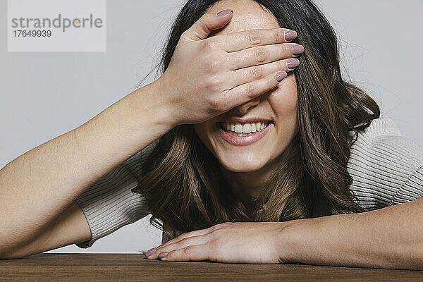 Happy woman covering eyes with hand at table against gray background