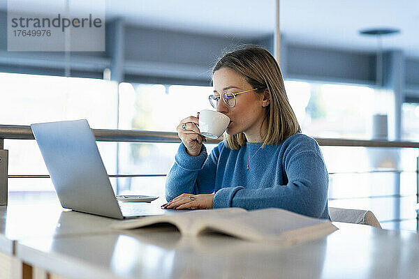 Junge Frau trinkt Kaffee und sitzt mit Laptop und Buch am Tisch