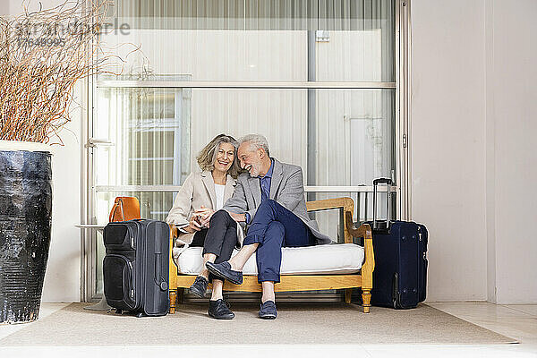 Fröhliches Seniorenpaar sitzt auf Bankgepäck vor Glas