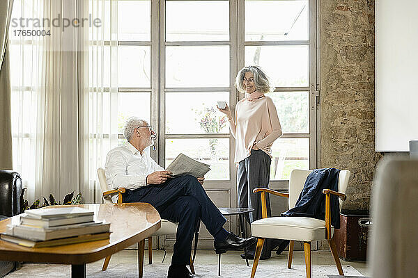 Lächelnde ältere Frau hält Kaffeetasse in der Hand und unterhält sich mit Mann im Wohnzimmer zu Hause