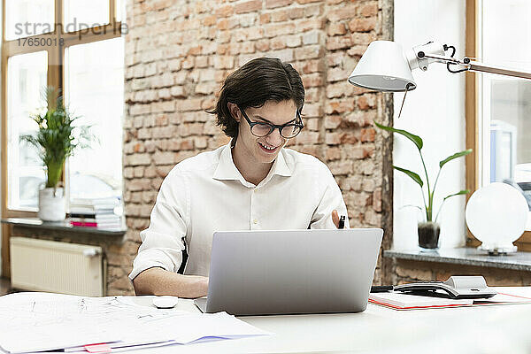 Glücklicher Geschäftsmann mit Laptop  der am Schreibtisch im Büro sitzt