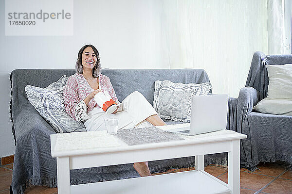 Fröhliche junge Frau mit Essen zum Mitnehmen sitzt zu Hause auf dem Sofa