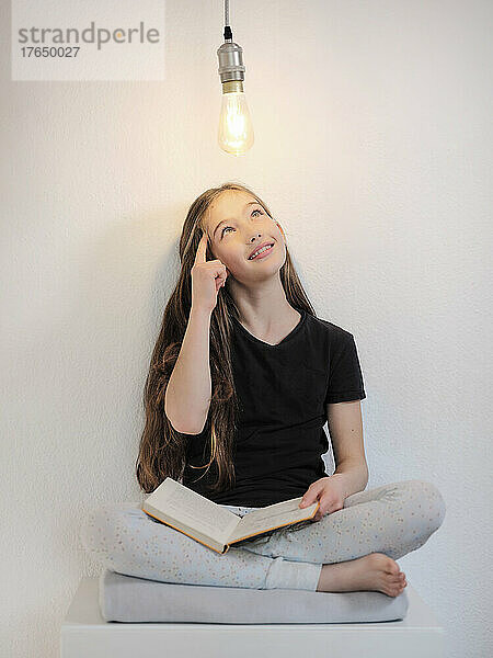 Nachdenkliches Mädchen mit Buch sitzt unter Glühbirne vor weißer Wand