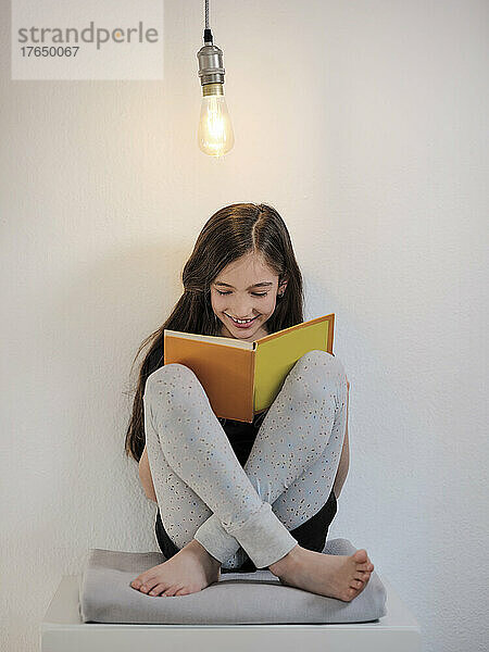 Glückliches Mädchen  das ein Buch liest und unter einer Glühbirne vor der Wand sitzt