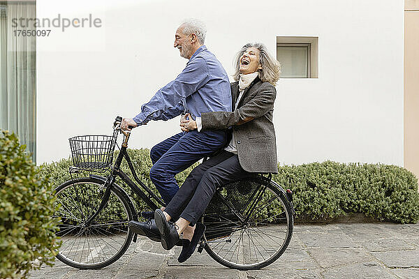 Glückliche ältere Frau sitzt mit Mann  der Fahrrad auf Fußweg fährt