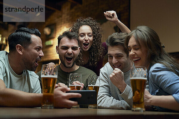 Gruppe fröhlicher Fußballfans  die in einer Kneipe Bier trinken und sich ein Spiel auf dem Smartphone ansehen