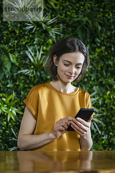 Lächelnde Frau benutzt Smartphone an Wand mit Pflanzen