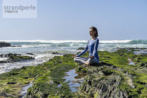 Frau sitzt im Lotussitz und meditiert auf einem Felsen am Strand