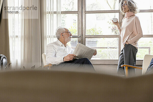 Lächelnde Frau hält eine Kaffeetasse in der Hand und unterhält sich mit einem Mann  der im heimischen Wohnzimmer eine Zeitung in der Hand hält