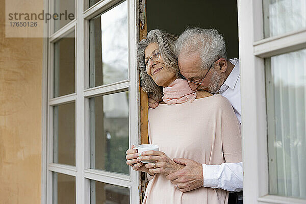 Affectionate senior man embracing woman holding coffee sup standing at window