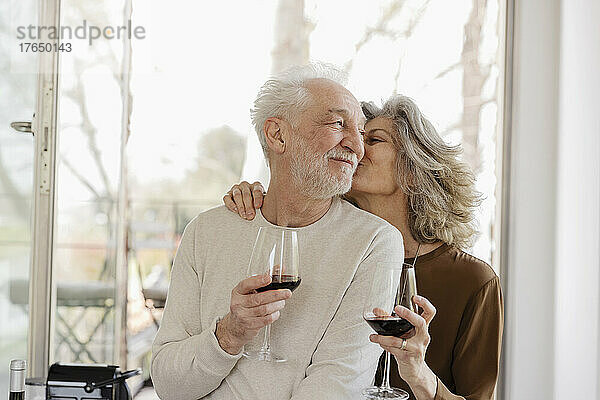 Ältere Frau hält Weinglas und küsst Mann vor Fenster in Hotelwohnung