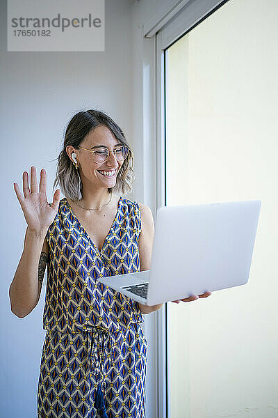 Glücklicher Freiberufler winkt bei einem Videoanruf über den Laptop am Fenster zu Hause mit der Hand