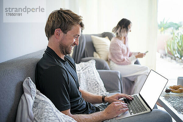 Glücklicher Freiberufler  der am Laptop arbeitet  während eine Frau ihr Smartphone im Wohnzimmer nutzt