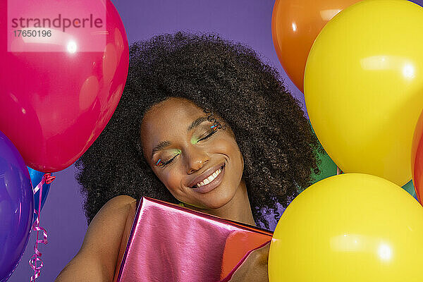 Fröhliche junge Frau mit geschlossenen Augen hält eine Geschenkbox in der Hand und steht inmitten bunter Luftballons vor violettem Hintergrund
