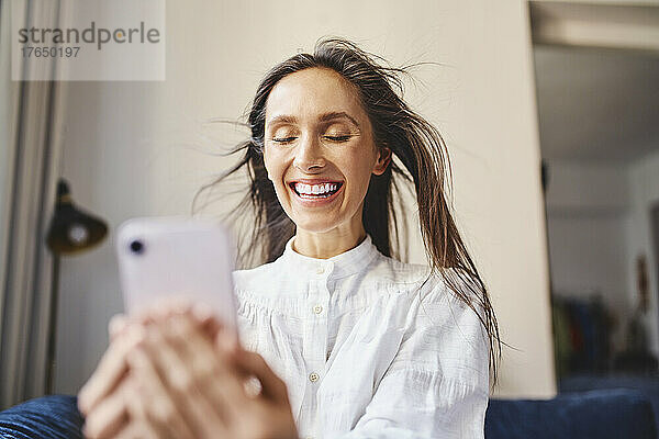 Glückliche Frau mit zerzausten Haaren hält ihr Smartphone zu Hause in der Hand