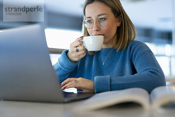 Junge Frau trinkt Kaffee und benutzt Laptop am Tisch