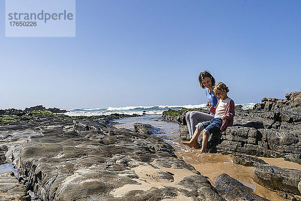 Glückliche Mutter und Tochter sitzen auf einem Felsen am sonnigen Strand
