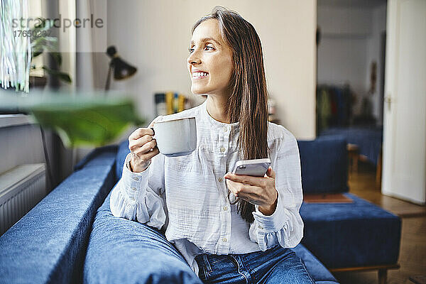 Lächelnde Frau mit Kaffeetasse und Smartphone sitzt zu Hause auf dem Sofa