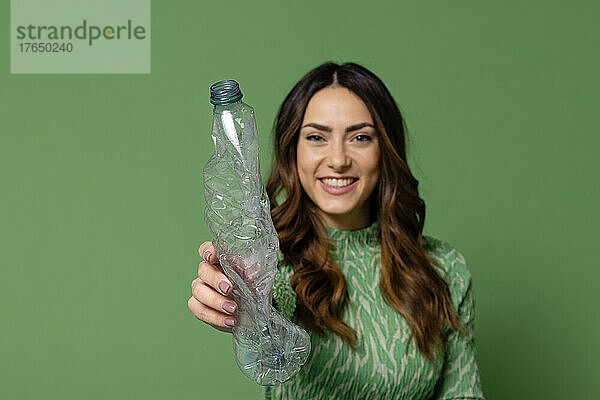 Lächelnde junge Frau zeigt Plastikflasche vor grünem Hintergrund