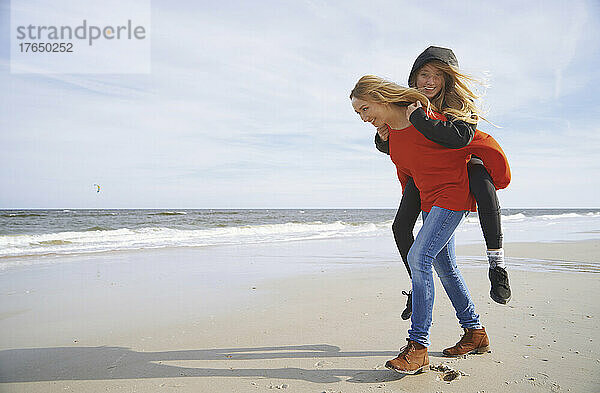 Glückliche blonde Mutter gibt ihrer Tochter an einem sonnigen Tag Huckepackfahrt am Strand