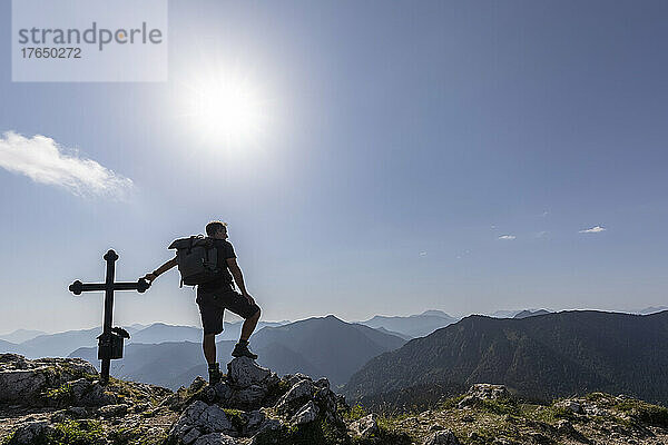Deutschland  Bayern  Sonne scheint über der Silhouette eines männlichen Wanderers  der am Gipfelkreuz des Fockensteins steht