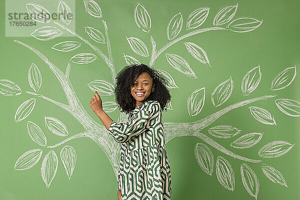 Lächelnde Frau steht vor einem Baum und zeichnet auf eine grüne Wand