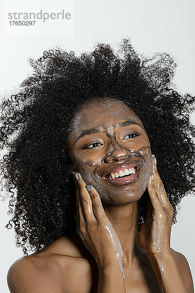 Glückliche junge Frau  die vor weißem Hintergrund eine Peeling-Gesichtsmaske auf ihr Gesicht aufträgt