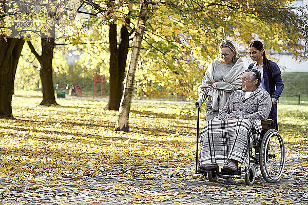 Hausmeister schiebt Rollstuhl eines behinderten Mannes von Frau  die mit Stock im Park geht
