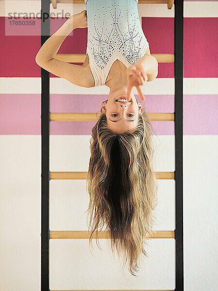 Smiling girl hanging upside down on wall bars gesturing peace sign