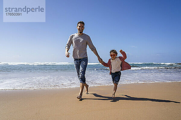 Glücklicher Vater und Tochter halten sich an den Händen und laufen an einem sonnigen Tag am Strand