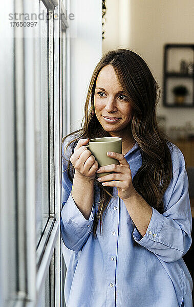Lächelnde schöne Frau mit Kaffeetasse  die zu Hause am Fenster steht