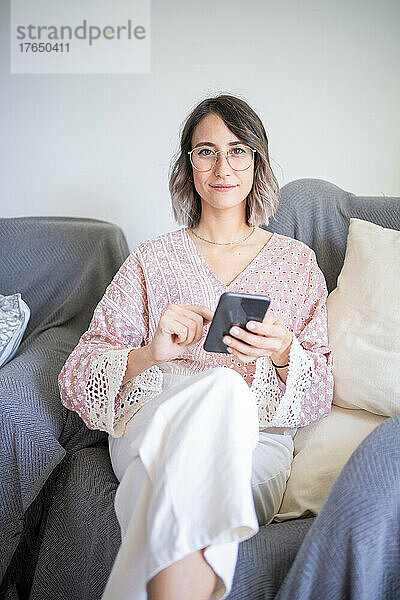 Junge Frau mit Smartphone sitzt zu Hause auf dem Sofa