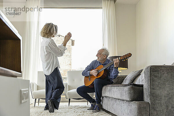 Älterer Mann spielt Gitarre und schaut Frau zu  die zu Hause im Wohnzimmer tanzt
