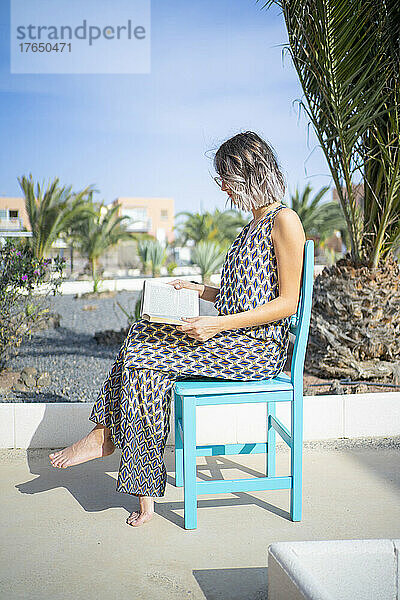 Junge Frau liest Buch und sitzt an einem sonnigen Tag auf einem blauen Stuhl