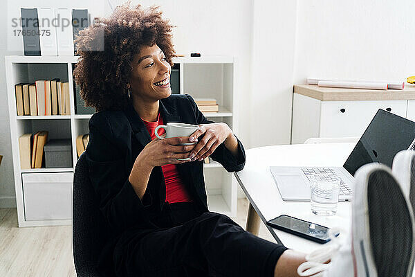 Lächelnde Geschäftsfrau sitzt mit Kaffeetasse am Schreibtisch im Büro