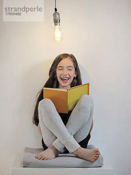 Cheerful girl with book sitting below light bulb in front of wall