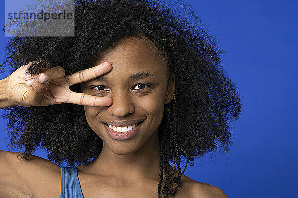 Lächelnde Frau gestikuliert vor blauem Hintergrund ein Friedenszeichen vor den Augen