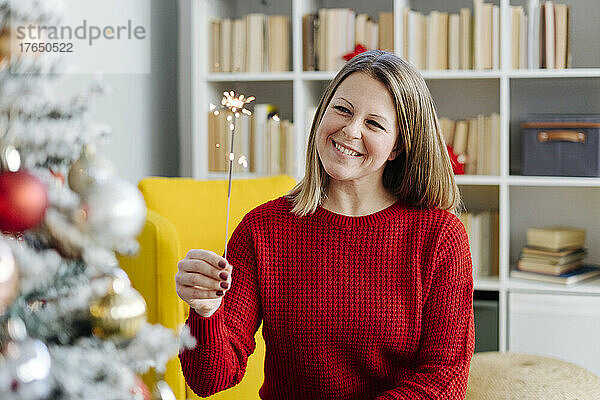 Glückliche Frau mit Wunderkerze sitzt zu Hause vor dem Weihnachtsbaum