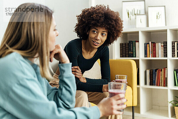 Junge Frau hält ein Glas Smoothie in der Hand und unterhält sich mit einer Freundin  die im Wohnzimmer sitzt