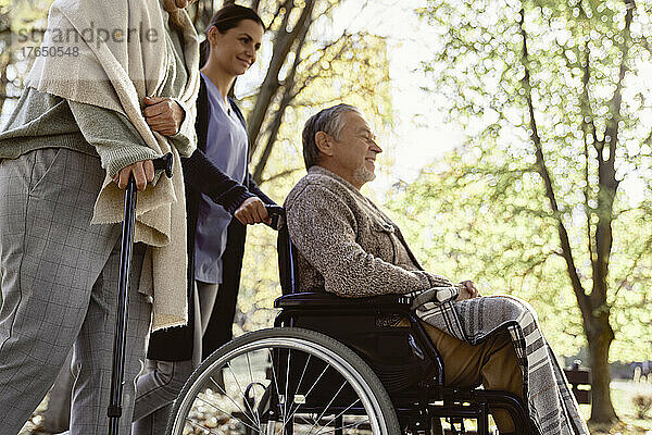 Ältere Frau geht mit Stock an Krankenschwester vorbei und behinderter Mann sitzt im Rollstuhl im Park