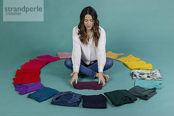 Young woman folding purple t-shirt sitting cross-legged amidst clothes against blue background