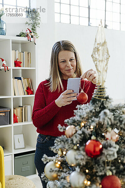 Lächelnde Frau fotografiert Christbaumkugel mit Handy vor dem heimischen Weihnachtsbaum