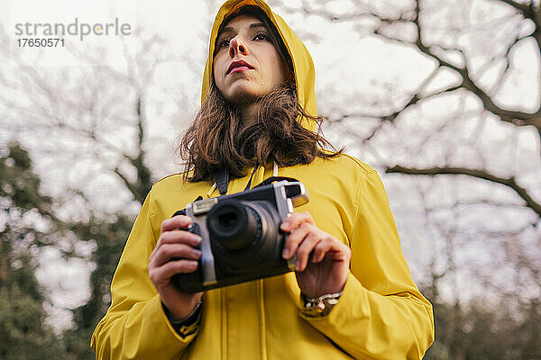 Junge Frau im gelben Regenmantel steht mit Kamera im Wald