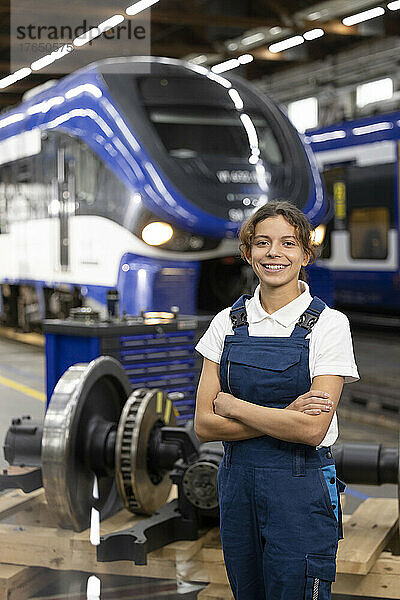 Lächelnder junger Auszubildender steht mit verschränkten Armen im Lager der Industrie