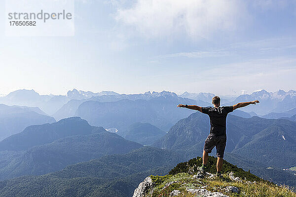 Mann steht an einem sonnigen Tag mit ausgestreckten Armen auf dem Berggipfel