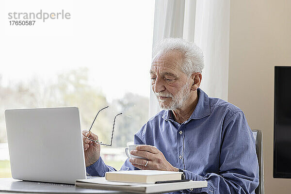 Älterer Mann mit Laptop und Kaffeetasse sitzt am Fenster einer Hotelwohnung