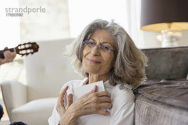 Lächelnde ältere Frau mit Brille sitzt mit Buch zu Hause auf dem Sofa
