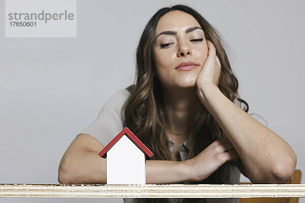 Lächelnde Frau mit Kopf in der Hand  die das Modell eines Hauses vor grauem Hintergrund betrachtet