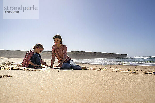 Glückliche Mutter und Tochter spielen an einem sonnigen Tag mit Sand am Strand