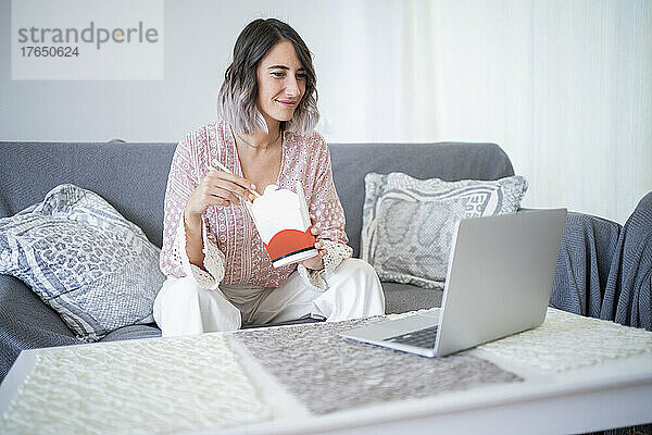Lächelnde Frau mit Essen zum Mitnehmen  die sich zu Hause auf dem Sofa ein Video über den Laptop ansieht